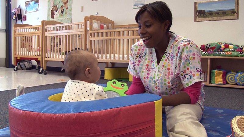 A caregiver sits and smiles with an infant